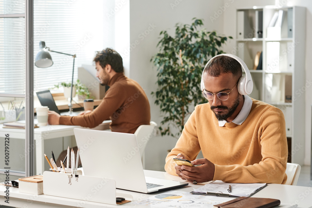 Serious young middle eastern manager in wireless headphones sitting at desk with laptop and texting sms in office Serious young middle eastern manager in wireless headphones sitting at desk with laptop and texting sms in office