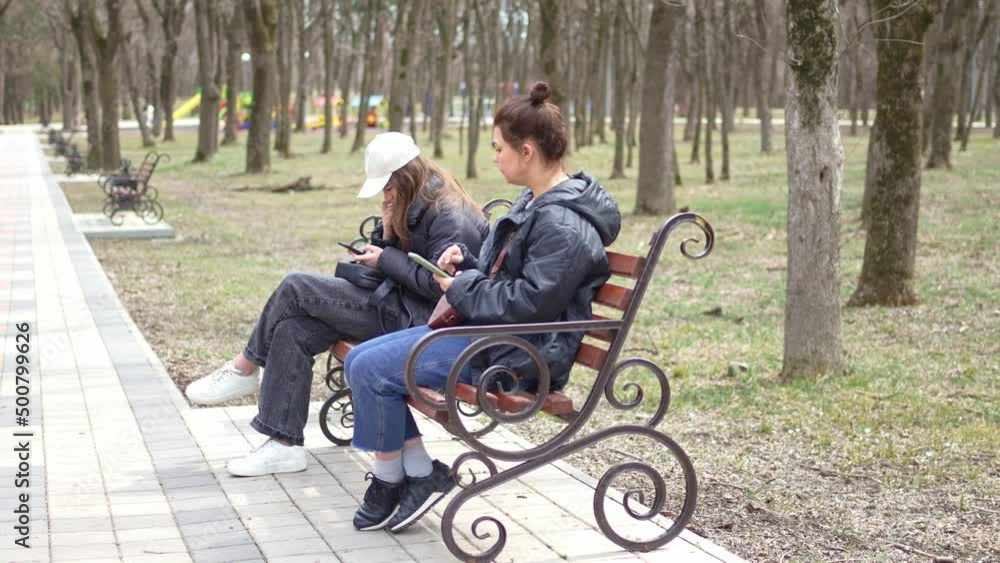 a woman and a girl in black jackets sit on a bench with smartphones. a woman and a girl in black jackets sit on a bench with smartphones.