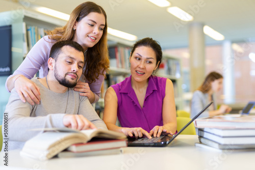 Group of enthusiastic young adult female and male colleagues working together in public library, discussing while sitting at table with laptop and books..