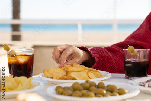 Close up shot of a hand picking up a potato chip at a table with snacks and drinks. Snack meal in a terrace in front of the beach. Two vermouths and food like crisps and olives.