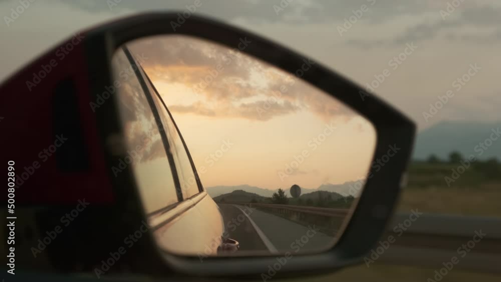 Side mirror of car driving fast reflects orange sunset and cloudy sky. Reflection of fields and trees on roadside at bright sunset extreme closeup Side mirror of car driving fast reflects orange sunset and cloudy sky. Reflection of fields and trees on roadside at bright sunset extreme closeup