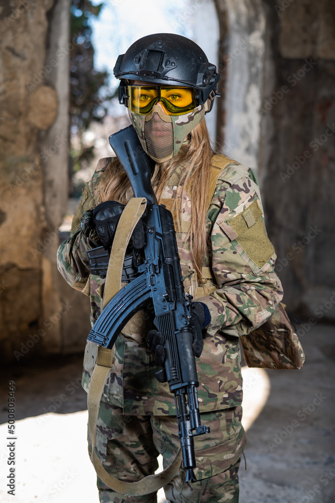 Fototapeta premium Portrait of a woman in a helmet and goggles with a machine gun in her hands. A female soldier in a camouflage uniform holds a weapon.