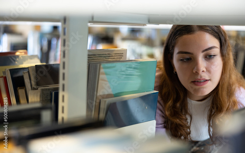 Young positive woman university student choosing books at library