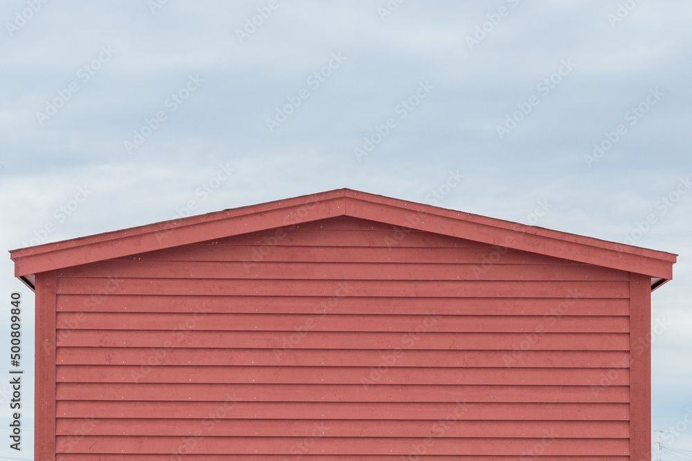 The exterior of a red wooden clapboard building with a peaked roof. The wood wall is horizontal cape cod beveled clapboard siding. The background is a blue sky with white clouds. The exterior of a red wooden clapboard building with a peaked roof. The wood wall is horizontal cape cod beveled clapboard siding. The background is a blue sky with white clouds.