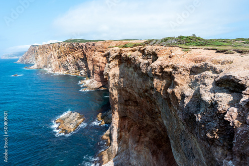 The horizon meets the bright blue sky and the deep blue ocean in the background. There's a steep cliff in the foreground with tundra like covering. There are waves at the bottom of the sea cliff. 