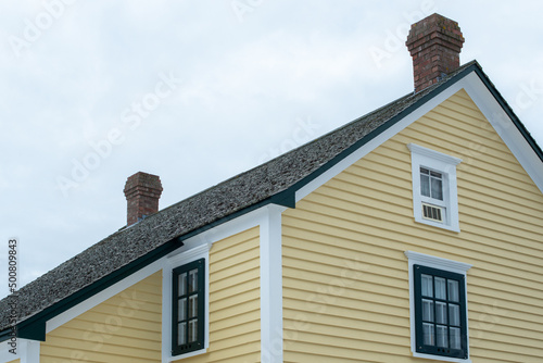 A large yellow wooden vintage building with lots of windows, dormers, and a peaked roof. The trim is white and black. There are two brick chimneys on the roof. The sky is grey and cloudy.