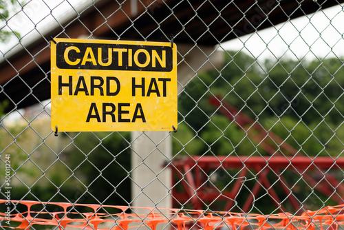 Yellow caution hard hat area sign on a chain link fence with a newly constructed bridge and crane in the background.
