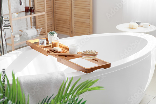 Wooden bathtub tray with different supplies and cosmetic products in light bathroom photo
