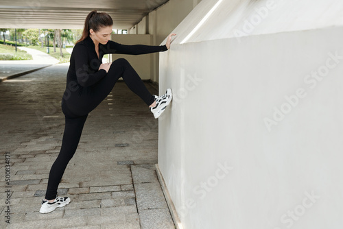 Fit serious young woman putting leg on wall to stretch after training