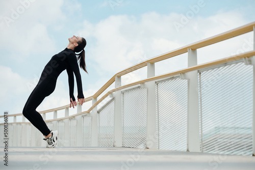 Happy female dancer in black jumpsuit doing toe stand on bridge