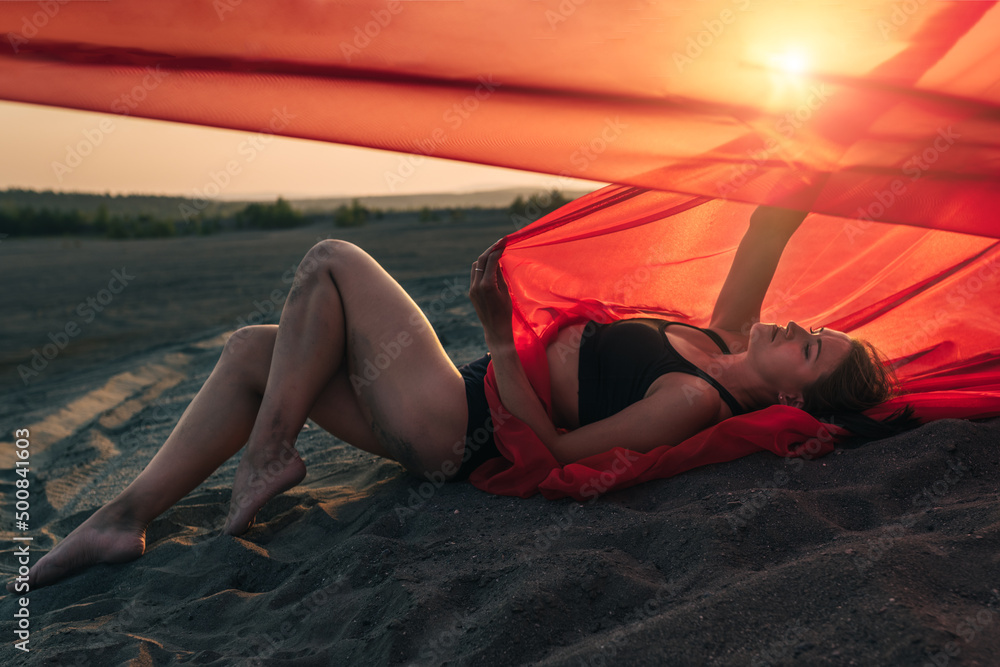 Elegant girl is on sand holding up long red fabric against sunset.