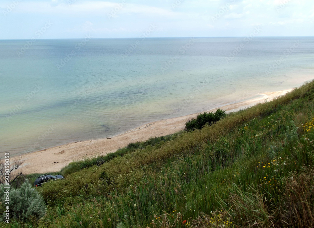 Azov sea coastal area with smooth green grass slope. Seashore view with sandy undeclared beach, blue sky horizon and a car.