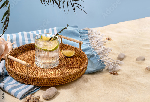 Fototapeta Naklejka Na Ścianę i Meble -  Gin tonic with lime on a wicker tray on beach with white sand. Summer sea vacation and travel concept. Exotic summer drinks.