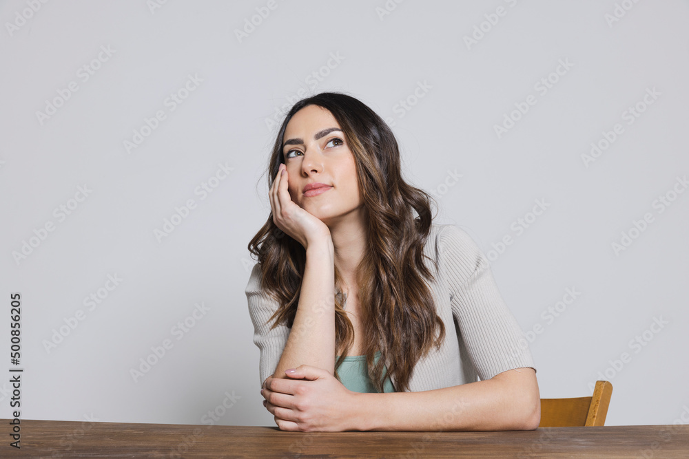 Thoughtful woman with head in hand sitting at table against gray background Thoughtful woman with head in hand sitting at table against gray background