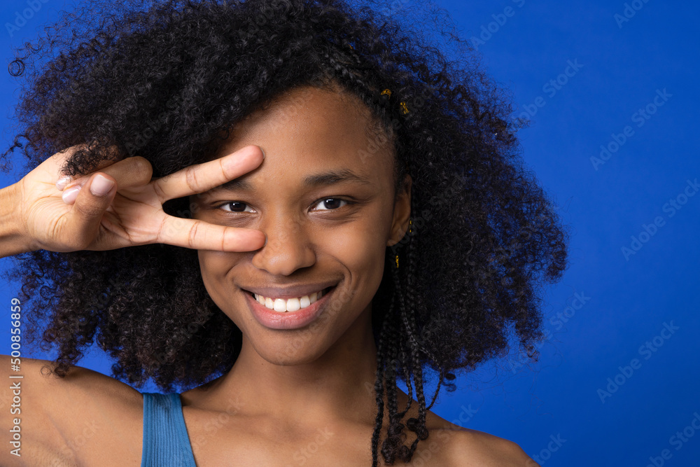 Smiling woman gesturing peace sign in front of eye against blue background Smiling woman gesturing peace sign in front of eye against blue background