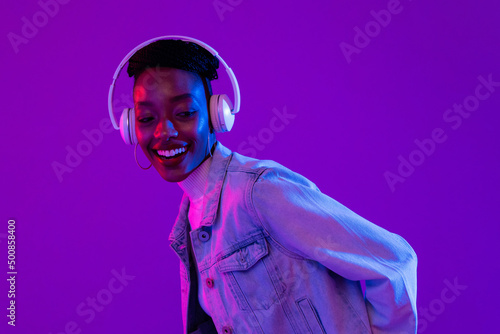 Trendy happy smiling African-American woman wearing headphones and listening to music in modern purple studio background