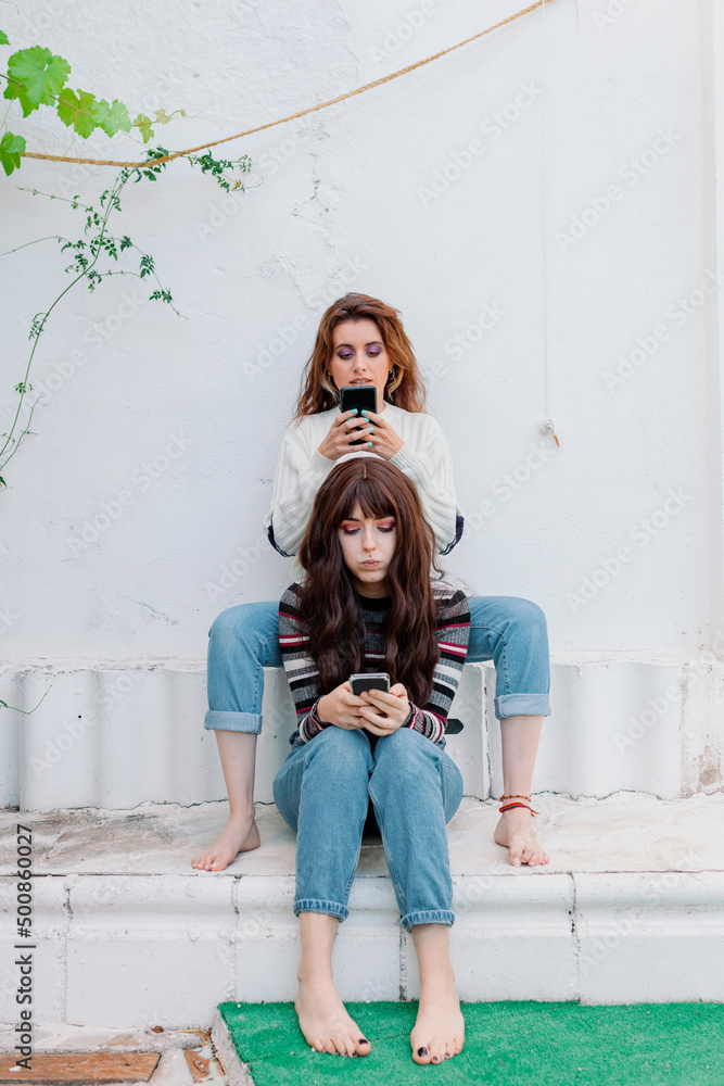 Friends with mobile phones sitting in front of white wall at back yard Friends with mobile phones sitting in front of white wall at back yard