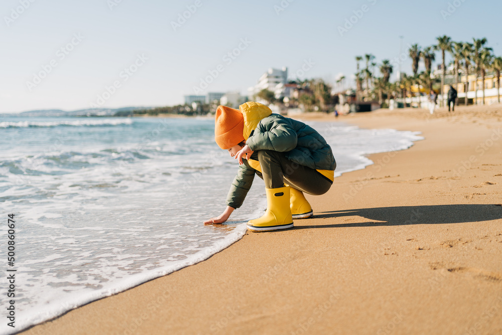 Obraz premium Boy in yellow rubber boots playing with water and sand at the beach. School kid touching water at autumn winter sea. Child having fun with waves at the shore. Spring Holiday vacation concept.