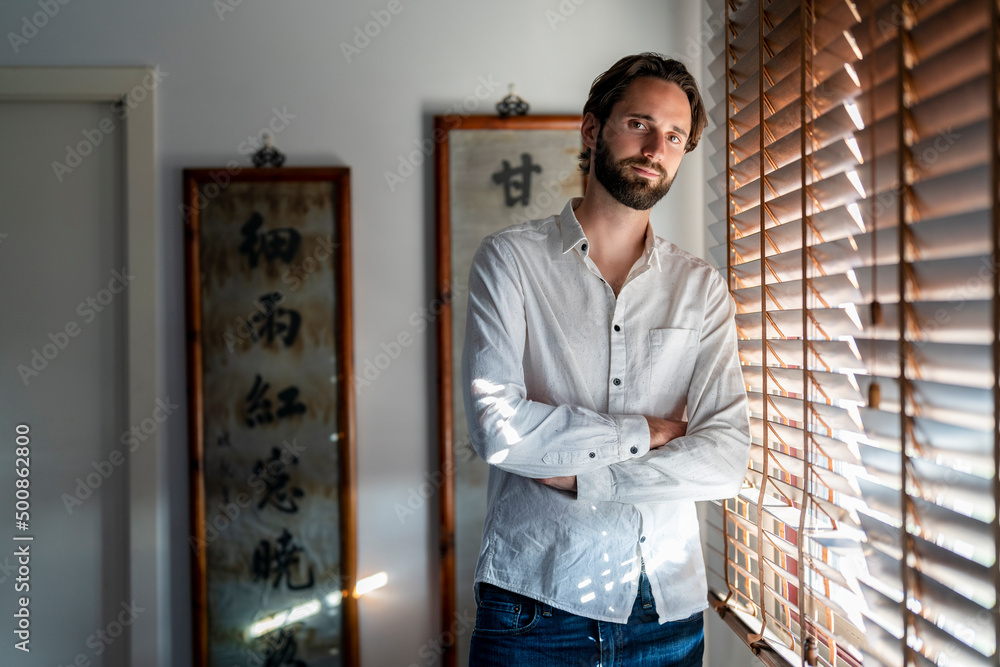 Portrait of man standing by blinded window with arms crossed Portrait of man standing by blinded window with arms crossed