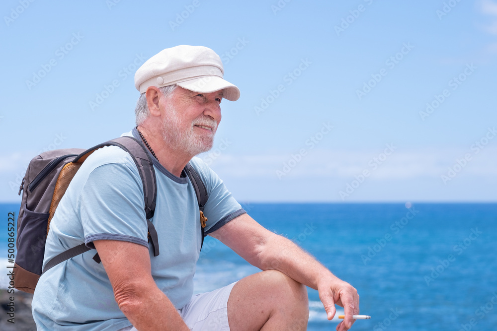 Attractive senior man with beard and hat sitting outdoors at sea looking at horizon while smoke a cigarette relaxing on a sunny day. Copy space, horizon over water. Retirement freedom vacation concept