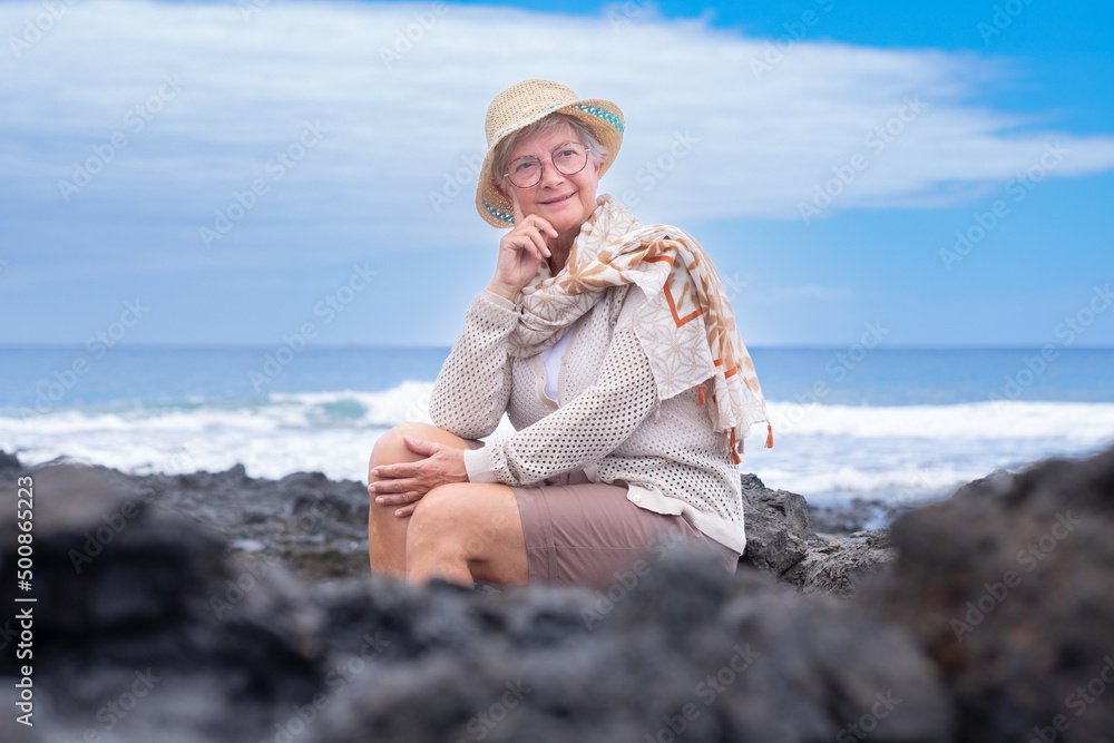 Portrait of smiling senior woman wearing eyeglasses and summer hat sitting outdoors at sea enjoying good time, freedom and relax. Cloudy sky, copy space. Travel vacation retirement concept
