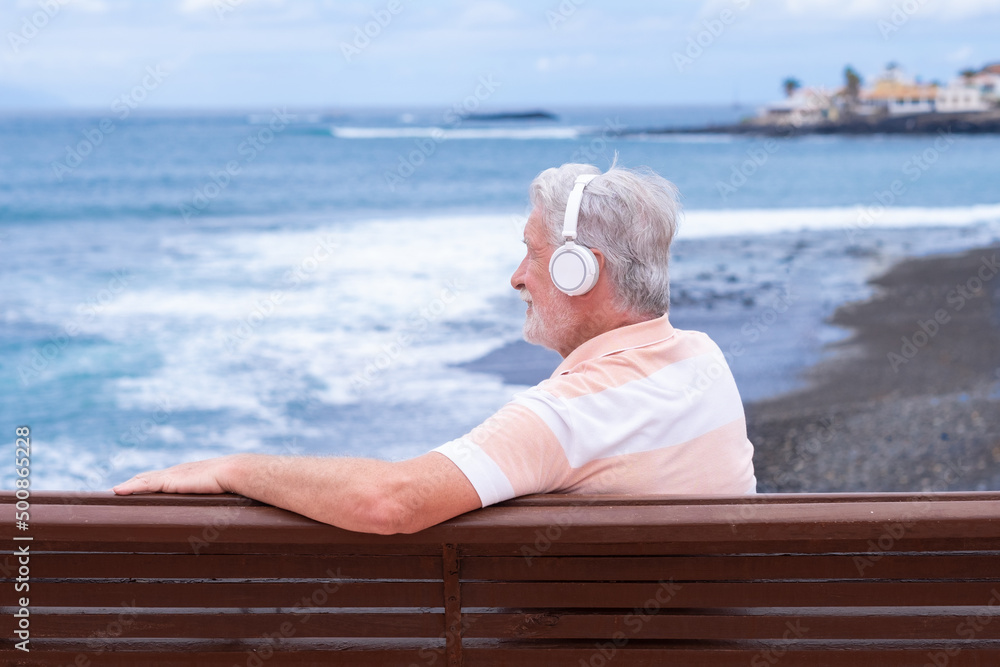 Senior bearded man wearing headphones sitting on outdoors bench at sea looking at horizon enjoying freedom and vacation. Active caucasian elderly man relaxing on a sunny day close to the beach