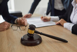 Close up of a judge's gavel on a wooden table, and an experienced lawyer working with notary documents and giving legal advice to a group of clients in the background. Law services concept