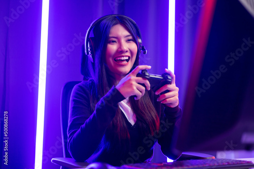 Playing video game. Young asian pretty woman sitting on chair holding joystick in living room. Happy female Professional Streamer chinese wearing headphone playing game in dark room neon light.