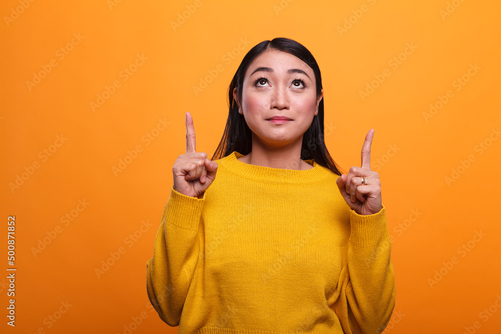 Friendly woman wearing yellow sweater looking up and pointing index fingers above head. Beautiful positive lady looking up and promoting sale offers and service business. Friendly woman wearing yellow sweater looking up and pointing index fingers above head. Beautiful positive lady looking up and promoting sale offers and service business.