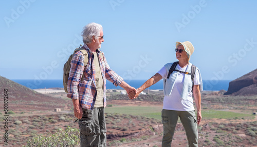 Happy active senior couple hiking in mountain enjoying healthy lifestyle and sunny day. Two elderly people hand in hand smiling, horizon over water. Vacation and sport concept