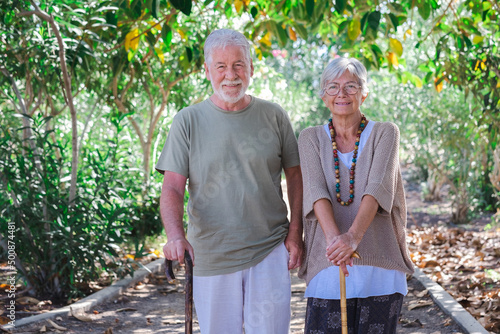 Beautiful white-haired senior couple walking in the woods with help of a walking cane. Smiling elderly grandparents enjoy healthy lifestyle in public park