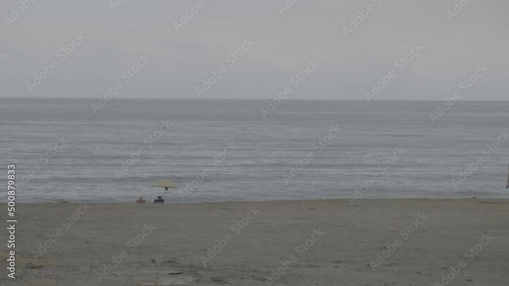 Two unrecognizable tourists on a beach under an umbrella looking at the sea on a cloudy day. Log Ungraded Two unrecognizable tourists on a beach under an umbrella looking at the sea on a cloudy day. Log Ungraded