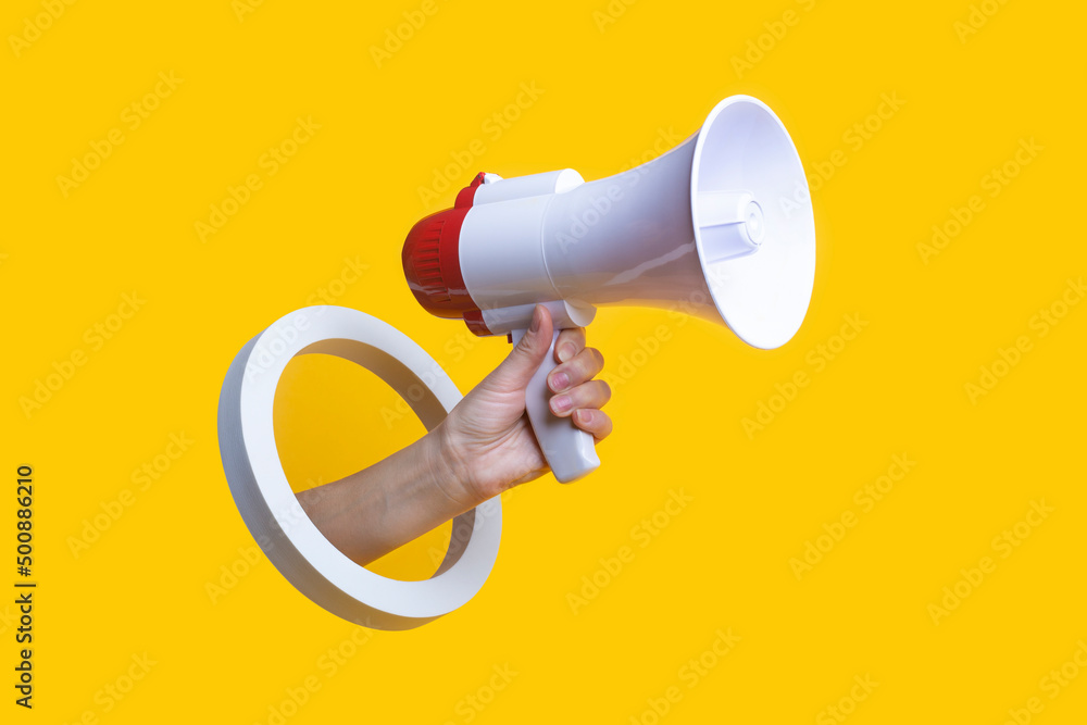 Female hand holds a megaphone in a round hole on a yellow background. - obrazy, fototapety, plakaty 