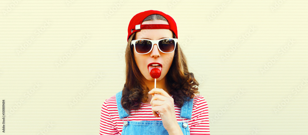 Portrait of young woman with lollipop wearing red baseball cap on white background