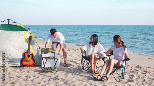 Happy young people enjoying with music at the beach, young man playing guitar and singing as his friends clapping and laughing, having fun on the beach picnic in summer holidays, vacation.