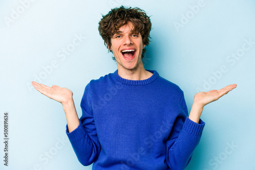 Young caucasian man isolated on blue background makes scale with arms, feels happy and confident.