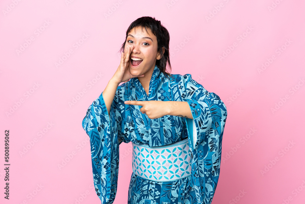 Young woman wearing kimono over isolated blue background pointing to the side to present a product and whispering something Young woman wearing kimono over isolated blue background pointing to the side to present a product and whispering something