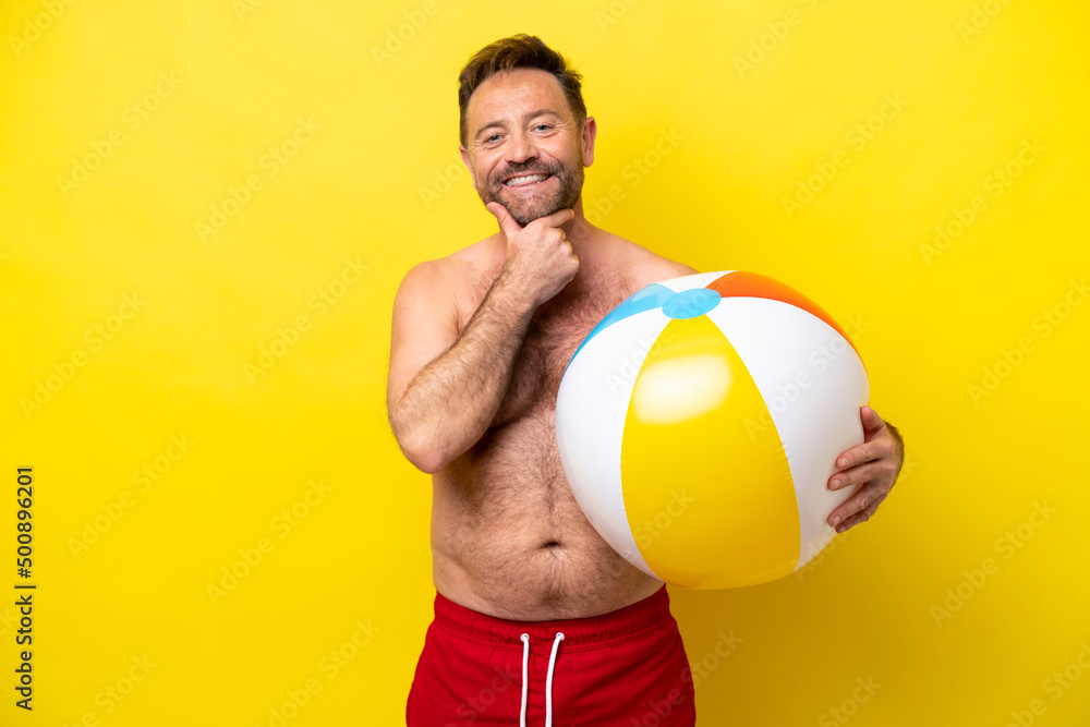 Middle age caucasian man holding beach ball isolated on yellow background happy and smiling