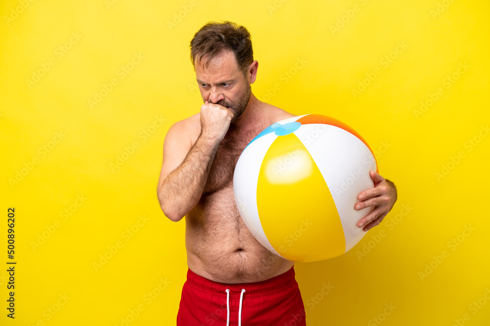 Middle age caucasian man holding beach ball isolated on yellow background having doubts