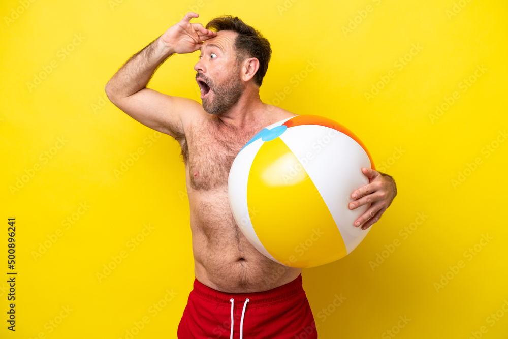 Middle age caucasian man holding beach ball isolated on yellow background doing surprise gesture while looking to the side