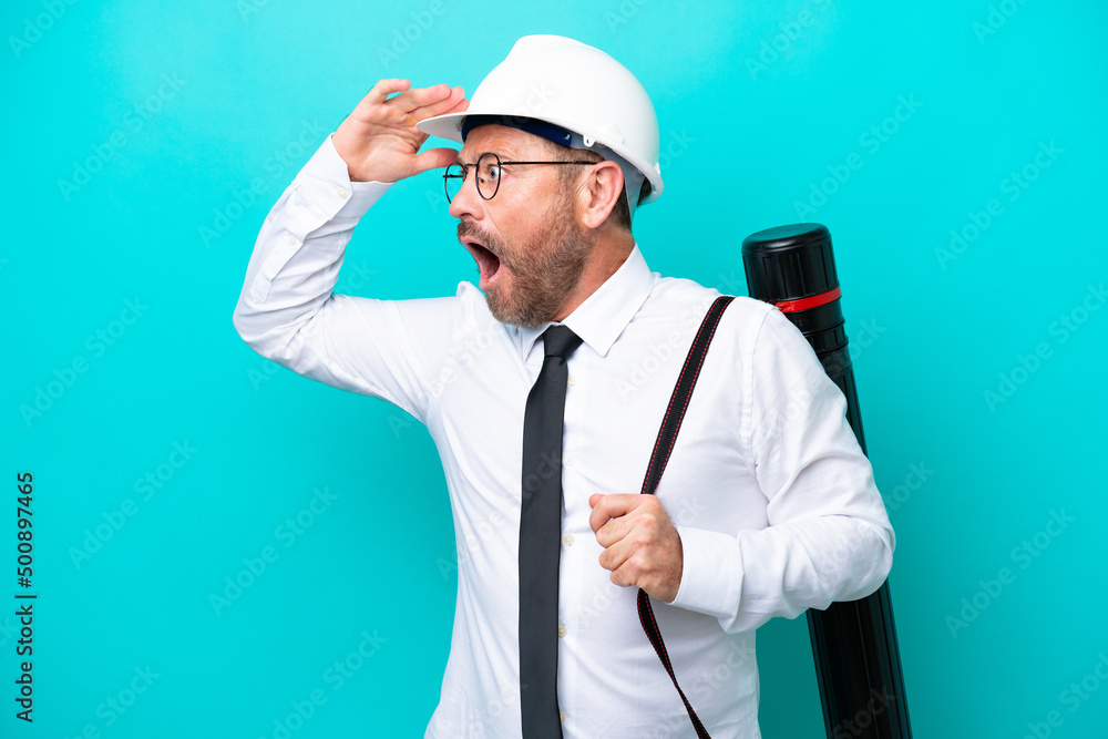 Middle age architect man with helmet and holding blueprints isolated on blue background doing surprise gesture while looking to the side Middle age architect man with helmet and holding blueprints isolated on blue background doing surprise gesture while looking to the side