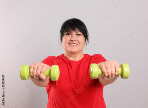 Happy overweight mature woman doing exercise with dumbbells on grey background