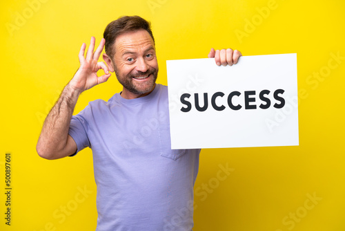 Middle age caucasian man isolated on yellow background holding a placard with text SUCCESS and celebrating a victory © luismolinero