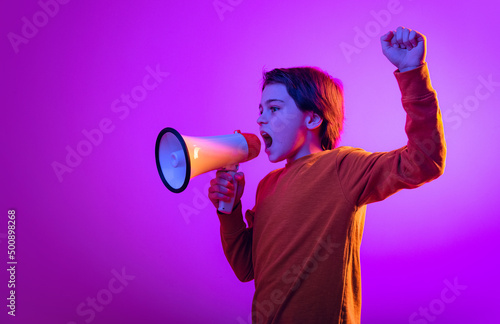 Portrait of boy, child shouting in megaphone, posing isolated over purple background in neon light
