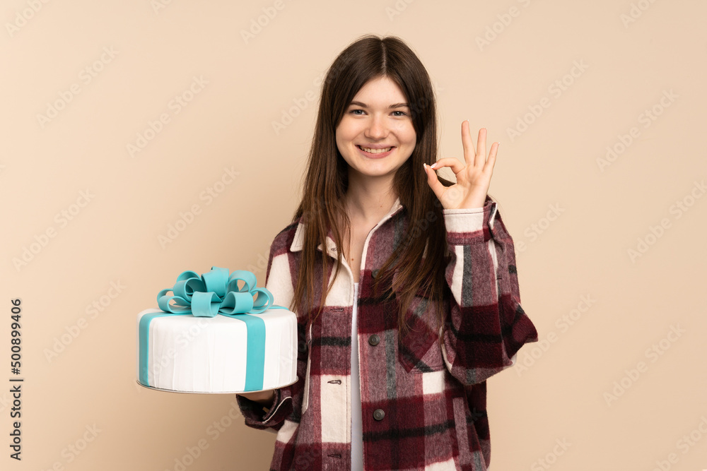 Young Ukrainian girl holding a big cake isolated on beige background showing ok sign with fingers Young Ukrainian girl holding a big cake isolated on beige background showing ok sign with fingers