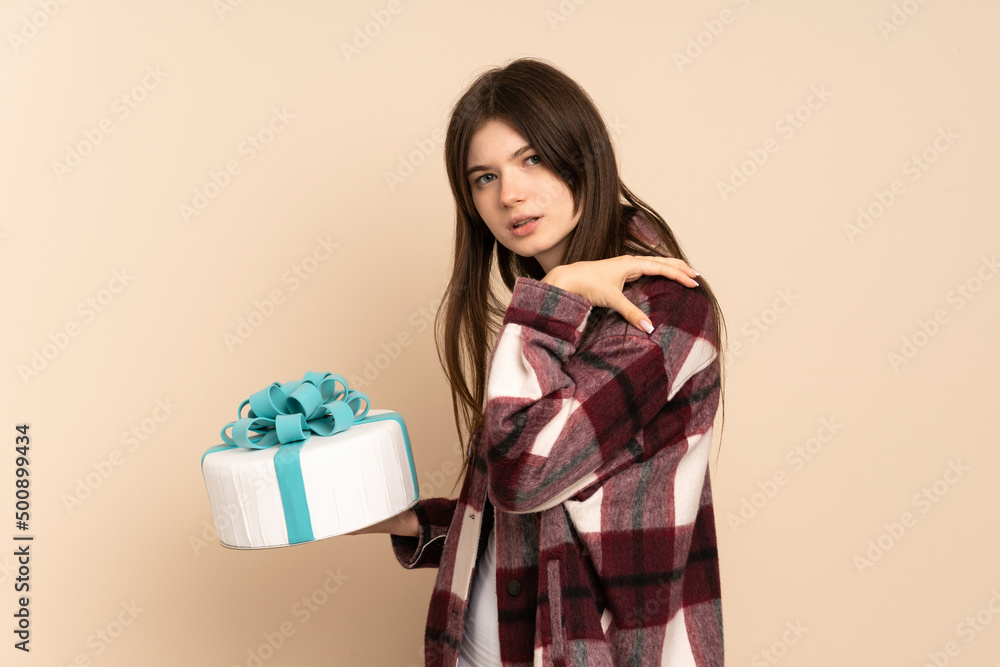 Young Ukrainian girl holding a big cake isolated on beige background suffering from pain in shoulder for having made an effort Young Ukrainian girl holding a big cake isolated on beige background suffering from pain in shoulder for having made an effort