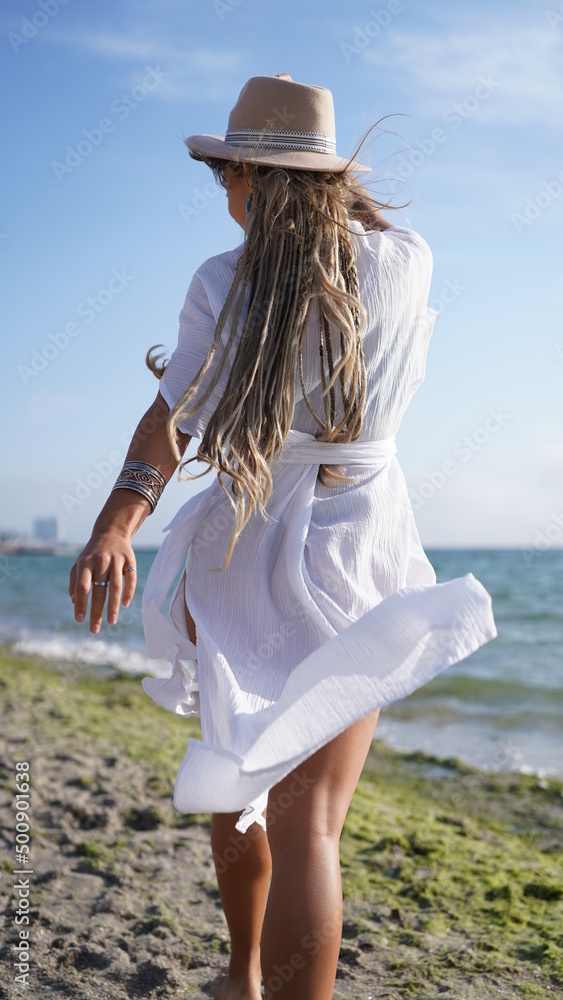 Back view of a girl with long dreadlocks in boho style. Charming girl in a hat walks along the beach near the ocean. Woman in white bohemian dress and jewelry on the beach. Hippie with tattoos travels