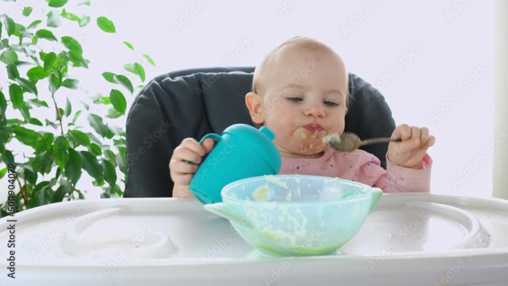 Baby learning to eat. Cute messy little girl eating with spoon and drinking by yourself in highchair. Baby led weaning method. Child 1 year old sitting in high chair, holding water bottle by her hand. Baby learning to eat. Cute messy little girl eating with spoon and drinking by yourself in highchair. Baby led weaning method. Child 1 year old sitting in high chair, holding water bottle by her hand.