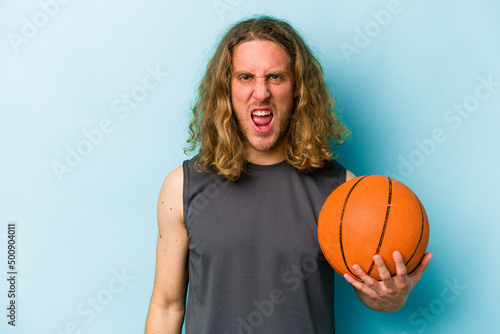 Young caucasian man playing basketball isolated on blue background screaming very angry and aggressive.