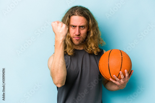 Young caucasian man playing basketball isolated on blue background showing fist to camera, aggressive facial expression.